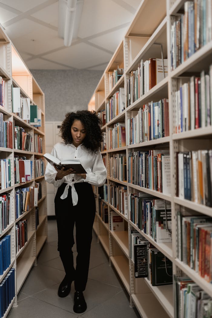 A woman with curly hair reading a book in a library aisle surrounded by shelves.